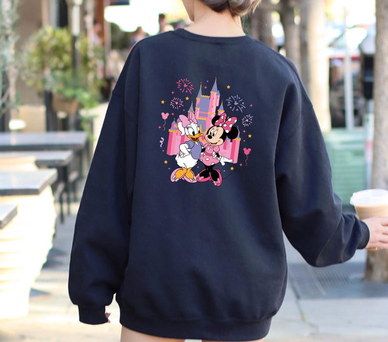 K&ouml;nnte beinhalten: Marineblaues Sweatshirt mit einem farbenfrohen Aufdruck von Daisy Duck und Minnie Mouse vor einem Schloss mit Feuerwerk. Die Grafik ist in Pink-, Lila- und Gelbt&ouml;nen gehalten. Das Sweatshirt hat lange &Auml;rmel.