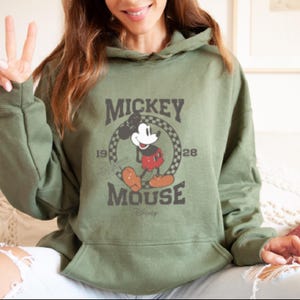 May include: A green hooded sweatshirt with a vintage Mickey Mouse graphic. The graphic features Mickey Mouse in a red and black outfit, standing in a circle with the text "Mickey Mouse" and the years "1928" and "1928".