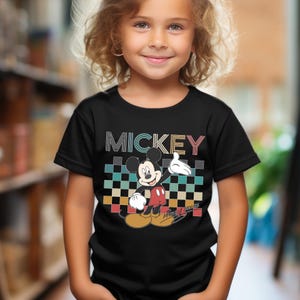 Puede incluir: Camiseta negra con un dise&ntilde;o de Mickey Mouse. El dise&ntilde;o incluye la palabra "MICKEY" en una fuente retro sobre un patr&oacute;n de cuadros. Mickey Mouse se representa con guantes blancos, pantalones cortos rojos y zapatos amarillos.