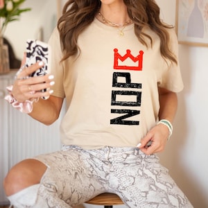 May include: A beige t-shirt with the word "NOPE" in black vertical letters, topped with a red crown. The model is wearing snake print pants and a gold necklace.