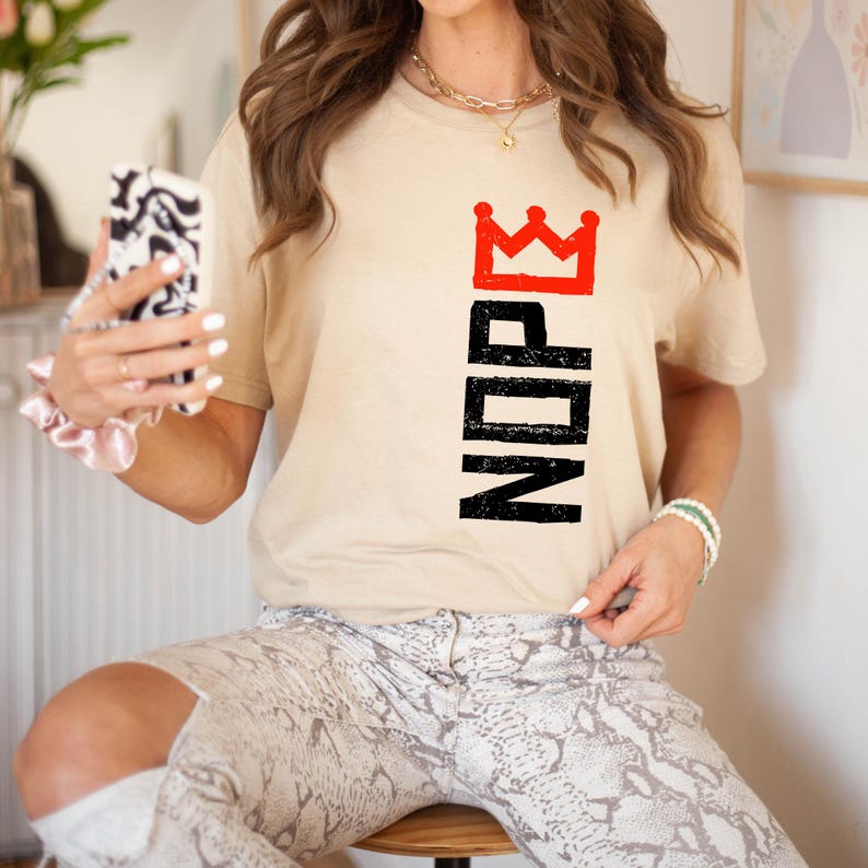 May include: A beige t-shirt with the word "NOPE" in black vertical letters, topped with a red crown. The model is wearing snake print pants and a gold necklace.