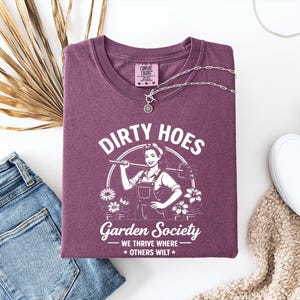 May include: A maroon t-shirt with the text "DIRTY HOES Garden Society" and a graphic of a woman with gardening tools. The shirt is folded with a silver necklace, blue jeans, and white slip-on shoes.