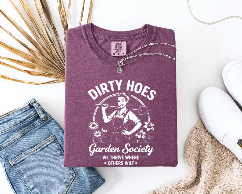 May include: A maroon t-shirt with the text "DIRTY HOES Garden Society" and a graphic of a woman with gardening tools. The shirt is folded with a silver necklace, blue jeans, and white slip-on shoes.