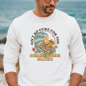 May include: White sweatshirt featuring a graphic of a skeleton surfing a wave with a guitar. The graphic is enclosed in a circle with the text "Ain't no cure for the summertime blues".