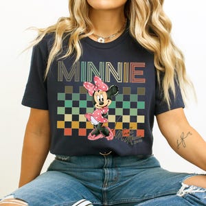 Puede incluir: Camiseta azul marino con un gr&aacute;fico de Minnie Mouse con un vestido de lunares rosa y un lazo. La palabra "MINNIE" est&aacute; impresa sobre un patr&oacute;n de cuadros. La camiseta tiene mangas cortas y cuello redondo. El dise&ntilde;o es retro.