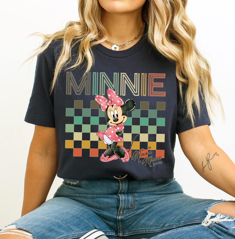 Puede incluir: Camiseta azul marino con un gr&aacute;fico de Minnie Mouse con un vestido de lunares rosa y un lazo. La palabra "MINNIE" est&aacute; impresa sobre un patr&oacute;n de cuadros. La camiseta tiene mangas cortas y cuello redondo. El dise&ntilde;o es retro.