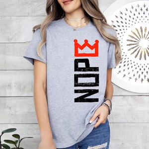 May include: A light gray t-shirt with the word "NOPE" in black vertical letters and a red crown graphic. The shirt is a casual style, suitable for everyday wear.