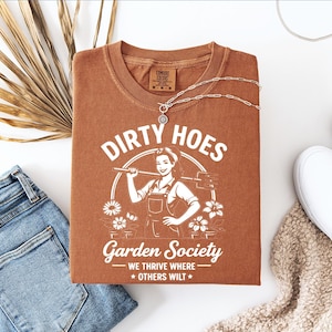 May include: A rust-colored t-shirt with the words "Dirty Hoes Garden Society" and a graphic of a woman holding a gardening tool. The shirt is paired with blue jeans, white slip-on shoes, and a silver necklace.