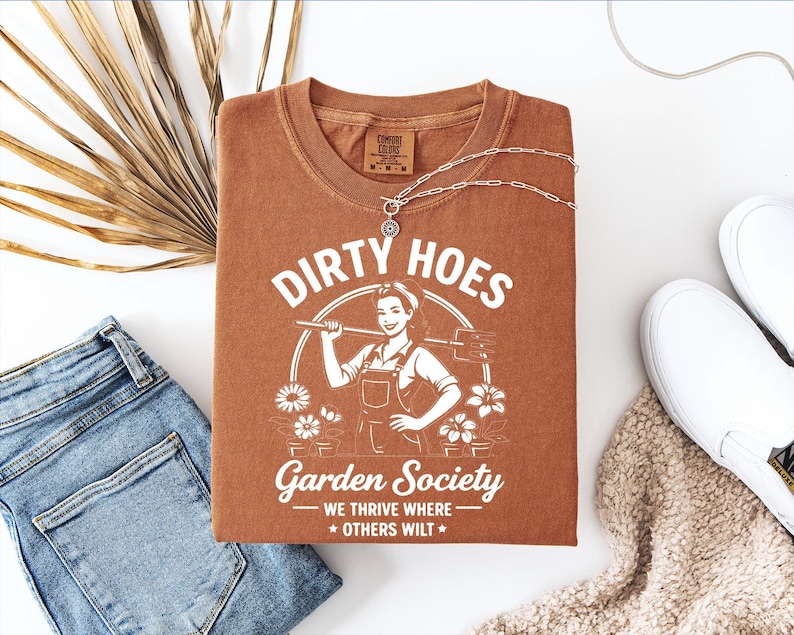 May include: A rust-colored t-shirt with the words "Dirty Hoes Garden Society" and a graphic of a woman holding a gardening tool. The shirt is paired with blue jeans, white slip-on shoes, and a silver necklace.