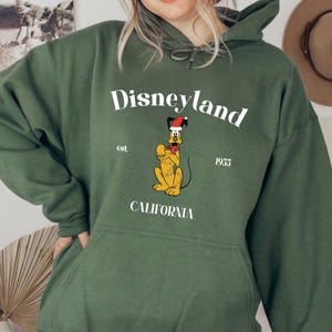 Op de afbeelding: Groen hoodie met een witte grafische print van Pluto met een kerstmanmuts. De tekst "Disneyland est. 1955 Californi&euml;" is onder de afbeelding gedrukt.