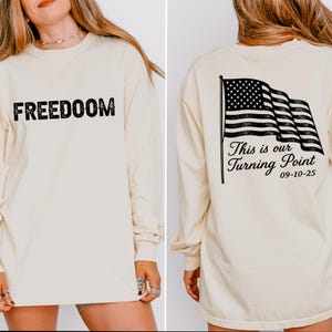 May include: A cream-coloured long-sleeved shirt with the word "FREEDOM" printed in a distressed black font on the front. The back displays a black and white American flag graphic with the text "This is our Turning Point" and the date "09-10-25".