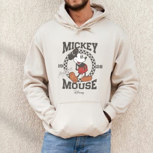 May include: A beige hoodie with a vintage Mickey Mouse graphic. The graphic features Mickey Mouse in a red and black outfit, standing in a checkered circle with the text "Mickey Mouse" and "1928" printed around the circle. The text "Disney" is printed below the graphic.