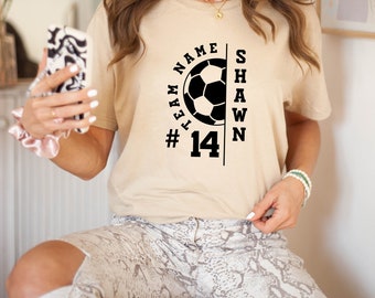 May include: A beige t-shirt with a black soccer ball graphic and the text "Team Name #14 Shawn" printed on the front.