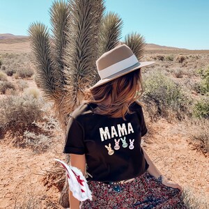 May include: A woman wearing a black t-shirt with the word "MAMA" and four colourful Easter bunny silhouettes. She is also wearing a brown hat with a white band, a brown belt, and a long floral skirt.