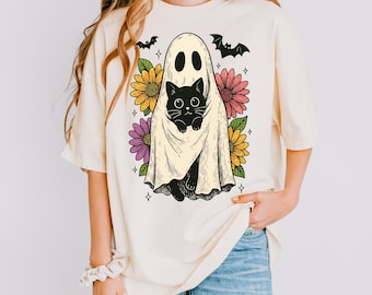 May include: Cream-colored t-shirt featuring a Halloween-themed graphic. The design includes a ghost holding a black cat, surrounded by colorful flowers, bats, and stars. The shirt is a casual, comfortable style.