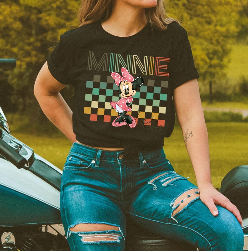 Puede incluir: Camiseta negra con la palabra "MINNIE" en una fuente retro sobre un gr&aacute;fico de Minnie Mouse. El gr&aacute;fico incluye un patr&oacute;n de cuadros en varios colores. Minnie Mouse lleva un lazo y un vestido rosa.