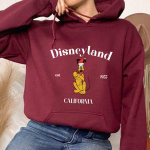 Op de afbeelding: Bordeauxkleurige hoodie met een witte afbeelding van Pluto met een kerstmanmuts en de tekst "Disneyland est. 1955 Californi&euml;".