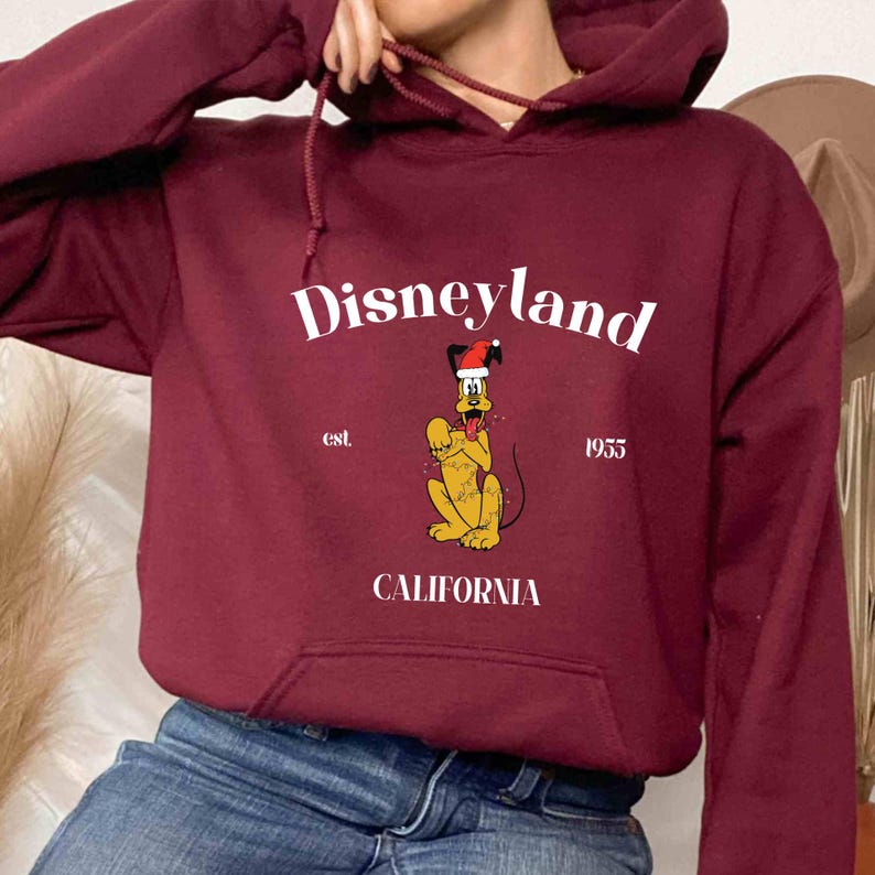 Op de afbeelding: Bordeauxkleurige hoodie met een witte afbeelding van Pluto met een kerstmanmuts en de tekst "Disneyland est. 1955 Californi&euml;".