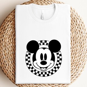 May include: A white t-shirt with a black and white checkered circle design featuring Mickey Mouse's face in the center.