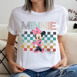 Puede incluir: Camiseta blanca con un dise&ntilde;o de Minnie Mouse. El dise&ntilde;o incluye la palabra "MINNIE" en una fuente a rayas sobre un patr&oacute;n de cuadros y una ilustraci&oacute;n de Minnie Mouse. La camiseta tambi&eacute;n tiene el texto "Minnie Mouse xoxo".
