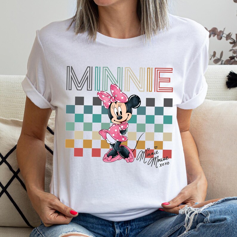 Puede incluir: Camiseta blanca con un dise&ntilde;o de Minnie Mouse. El dise&ntilde;o incluye la palabra "MINNIE" en una fuente a rayas sobre un patr&oacute;n de cuadros y una ilustraci&oacute;n de Minnie Mouse. La camiseta tambi&eacute;n tiene el texto "Minnie Mouse xoxo".