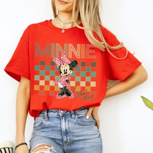 Puede incluir: Una camiseta roja con la palabra "MINNIE" en una fuente retro y un dibujo de Minnie Mouse. Minnie lleva un vestido y un lazo de lunares rosas. El fondo tiene un patr&oacute;n de cuadros en tonos rojos, verdes y marrones.