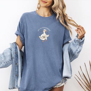 May include: A blue short-sleeved t-shirt featuring a graphic of a white goose wearing a lab coat and goggles, holding a beaker. The text "OFF TO EXPERIMENT" is printed in an arc above the goose. The shirt has a crew neckline and a relaxed fit.
