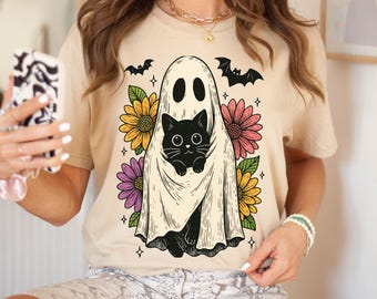 May include: Beige t-shirt featuring a Halloween-themed graphic. The design includes a ghost holding a black cat, surrounded by colorful flowers and bats. The shirt is a casual, short-sleeved style.