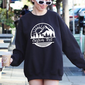 May include: Black sweatshirt with a white mountain scene graphic and the text "Custom Text" in a circle.