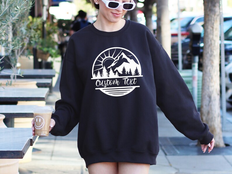 May include: Black sweatshirt with a white mountain scene graphic and the text "Custom Text" in a circle.