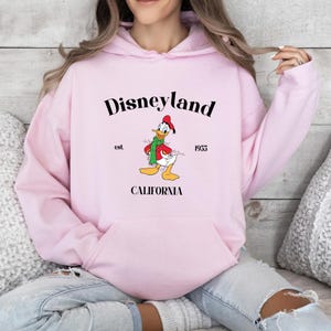 Op de afbeelding: Roze hoodie met een zwarte afbeelding van Donald Duck met een rode muts en een groene sjaal. De afbeelding bevat ook de tekst "Disneyland est. 1955 CALIFORNI&Euml;".