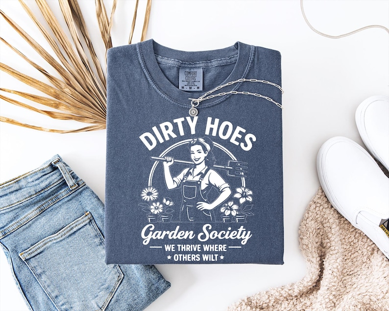 May include: A navy blue t-shirt with the text "DIRTY HOES Garden Society" and a graphic of a woman holding a gardening tool. The shirt is folded and displayed with a silver necklace, blue jeans, and white shoes.