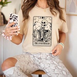 May include: A beige t-shirt featuring a black and white illustration of a skeleton sitting with its hands clasped. The illustration is surrounded by flowers and the text "THE OVERTHINKER".