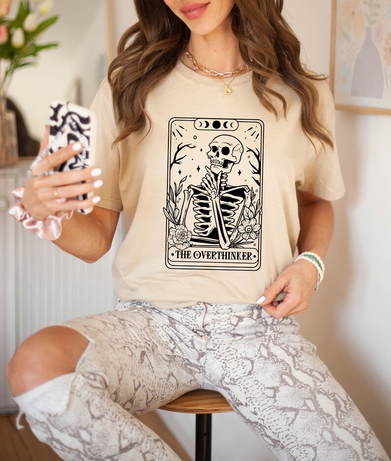May include: A beige t-shirt featuring a black and white illustration of a skeleton sitting with its hands clasped. The illustration is surrounded by flowers and the text "THE OVERTHINKER".