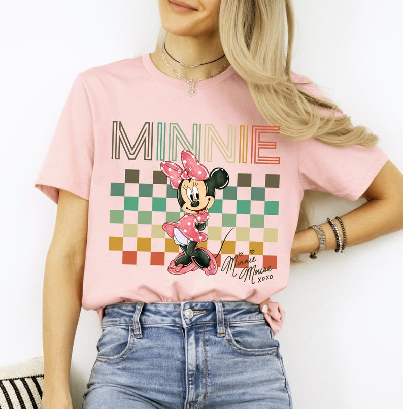 Puede incluir: Camiseta rosa claro con un gr&aacute;fico de Minnie Mouse. El dise&ntilde;o incluye la palabra "MINNIE" en una fuente retro sobre un patr&oacute;n de cuadros y una ilustraci&oacute;n de Minnie Mouse. La camiseta tambi&eacute;n tiene el texto "Minnie Mouse xoxo".