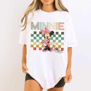 Puede incluir: Camiseta blanca con un gr&aacute;fico de Minnie Mouse con un vestido de lunares rosa y un lazo. La palabra "MINNIE" est&aacute; encima del personaje en una fuente retro, con un fondo a cuadros multicolor. La camiseta tambi&eacute;n tiene el texto "Minnie Mouse xoxo".