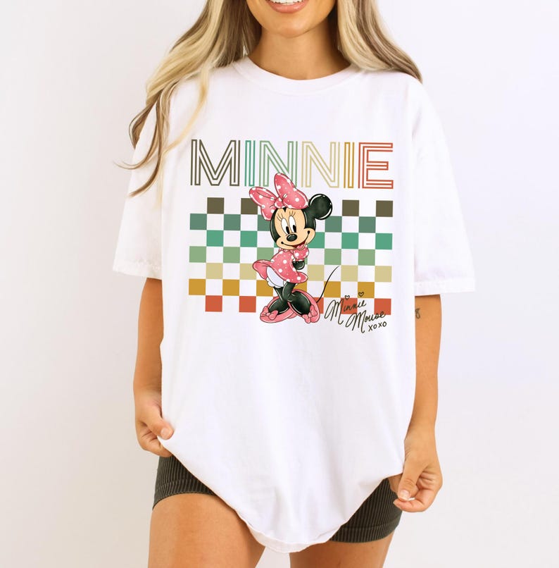 Puede incluir: Camiseta blanca con un gr&aacute;fico de Minnie Mouse con un vestido de lunares rosa y un lazo. La palabra "MINNIE" est&aacute; encima del personaje en una fuente retro, con un fondo a cuadros multicolor. La camiseta tambi&eacute;n tiene el texto "Minnie Mouse xoxo".