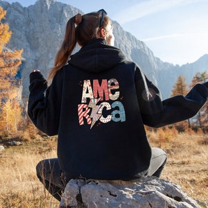 May include: Black hoodie featuring the word "America" in a patriotic design on the back. The letters are in red, white, and blue with a lightning bolt graphic. The hoodie is worn outdoors.