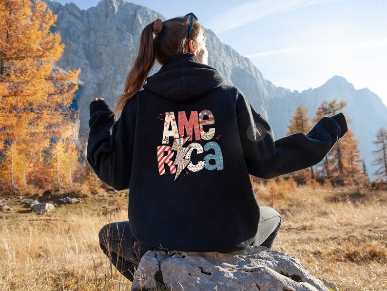 May include: Black hoodie featuring the word "America" in a patriotic design on the back. The letters are in red, white, and blue with a lightning bolt graphic. The hoodie is worn outdoors.