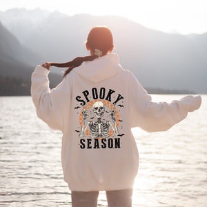 May include: White hoodie with a Halloween-themed graphic on the back. The design features a skeleton with peace signs, surrounded by bats and the words "Spooky Season" in a vintage font. The hoodie is a casual, comfortable style.