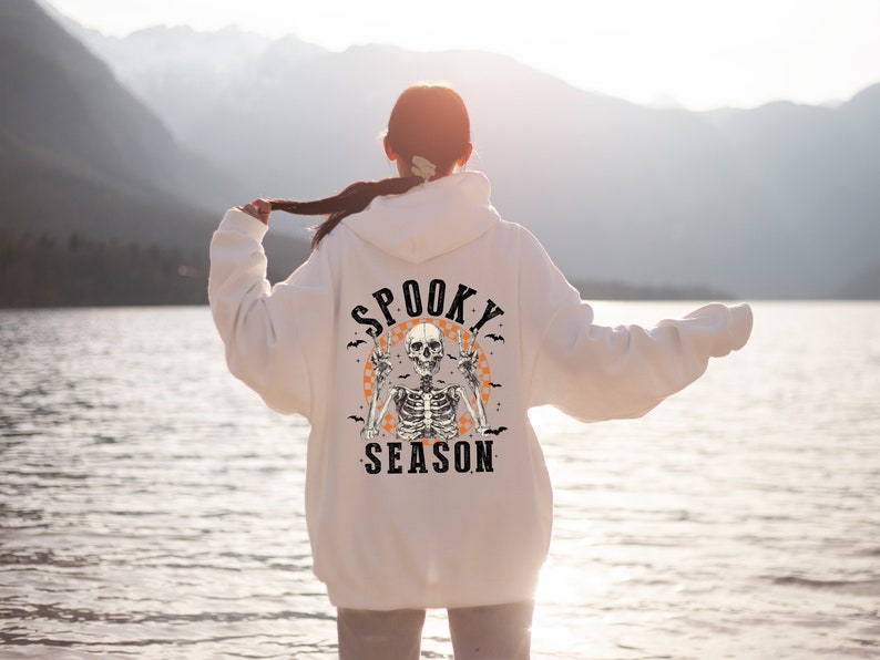 May include: White hoodie with a Halloween-themed graphic on the back. The design features a skeleton with peace signs, surrounded by bats and the words "Spooky Season" in a vintage font. The hoodie is a casual, comfortable style.