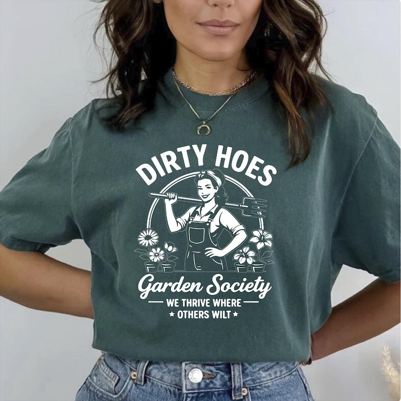 May include: Dark green t-shirt with the text "DIRTY HOES Garden Society" and a graphic of a woman holding a gardening tool. The shirt also features the phrase "WE THRIVE WHERE OTHERS WILT" below the main text.