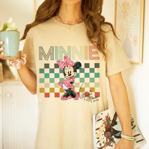 Puede incluir: Camiseta beige con un estampado de Minnie Mouse con vestido y lazo rosa de lunares. La palabra "MINNIE" est&aacute; impresa en la parte superior con una fuente retro, con un patr&oacute;n de cuadros debajo. La camiseta tiene mangas cortas.