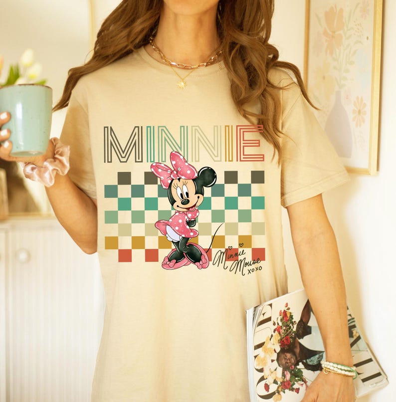 Puede incluir: Camiseta beige con un estampado de Minnie Mouse con vestido y lazo rosa de lunares. La palabra "MINNIE" est&aacute; impresa en la parte superior con una fuente retro, con un patr&oacute;n de cuadros debajo. La camiseta tiene mangas cortas.