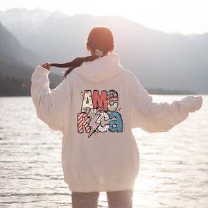 May include: White hoodie featuring the word "AMERICA" in a colourful, patriotic design on the back. The design includes stars, stripes, and a lightning bolt. The hoodie is a casual, comfortable style.