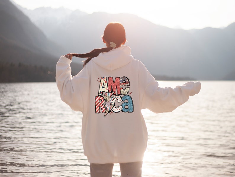 May include: White hoodie featuring the word "AMERICA" in a colourful, patriotic design on the back. The design includes stars, stripes, and a lightning bolt. The hoodie is a casual, comfortable style.