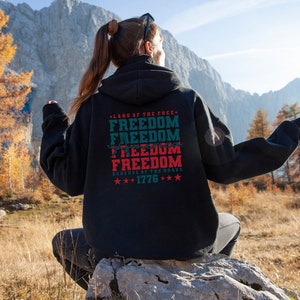 May include: Black hooded sweatshirt with a red, white and blue graphic print that says "Land of the Free, Freedom, Freedom, America, Freedom, Freedom, Because of the Brave, 1776".