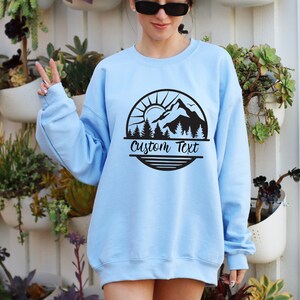 May include: Light blue sweatshirt with a black mountain scene graphic and the text "Custom Text" in a circle.