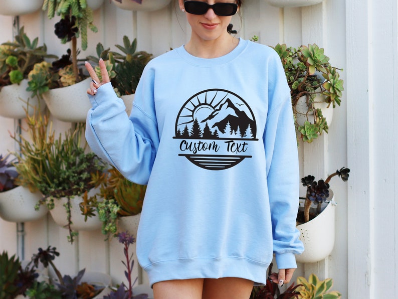 May include: Light blue sweatshirt with a black mountain scene graphic and the text "Custom Text" in a circle.