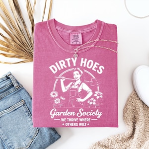 May include: A pink t-shirt with the words "Dirty Hoes Garden Society" and a graphic of a woman holding a gardening tool. The shirt is paired with blue jeans, white sneakers, and a necklace.